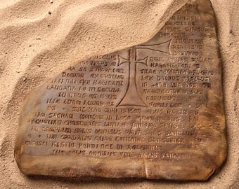 May include: A tan, irregularly shaped stone tablet featuring an engraved cross in the center and extensive Latin text inscribed across its surface. The tablet has a smooth, weathered texture and is partially embedded in fine, light-colored sand.