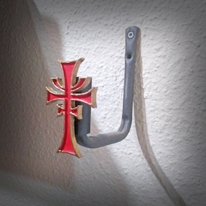May include: A black metal hook with a red and gold cross design. The hook is mounted on a white wall.