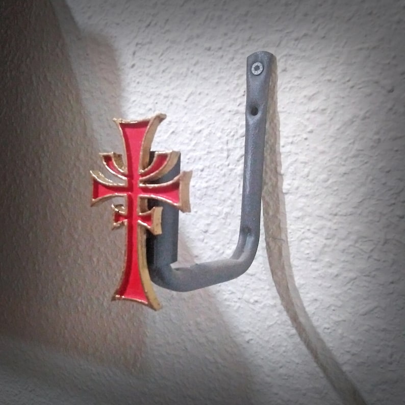 May include: A black metal hook with a red and gold cross design. The hook is mounted on a white wall.