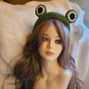 May include: A green crocheted frog headband with white eyes. The headband is worn on a mannequin with long brown hair.