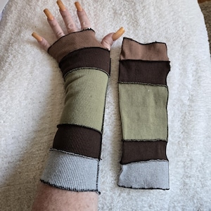 May include: A pair of fingerless arm warmers in shades of brown, olive green, and gray. The ribbed fabric is stitched together in horizontal stripes. The arm warmers are designed to cover the forearm and hand, leaving the fingers exposed.