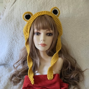 May include: A mustard yellow crochet hat featuring two prominent, rounded frog-like ears on top. The ears are detailed with white and black circular centers. The hat has long, dangling side panels that frame the face, crafted in a textured knit.