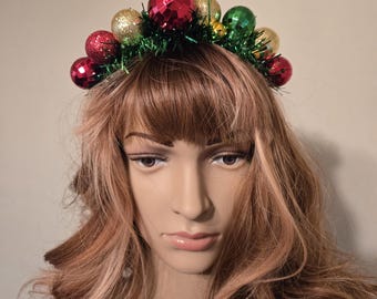 Diadema navideña verde con espumillón rojo, verde y plateado. Bolas y cascabeles navideños. Banda forrada de tela. Ideal para fiestas familiares o de trabajo.