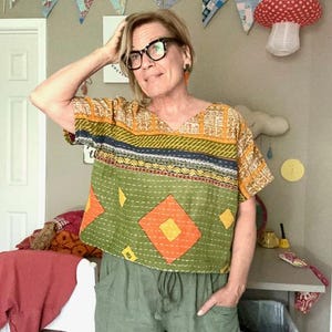 May include: A short-sleeved, boxy-fit top featuring a patchwork design with olive green, mustard yellow, and orange diamond patterns, accented by horizontal bands of dark blue and intricate white stitching throughout the fabric.