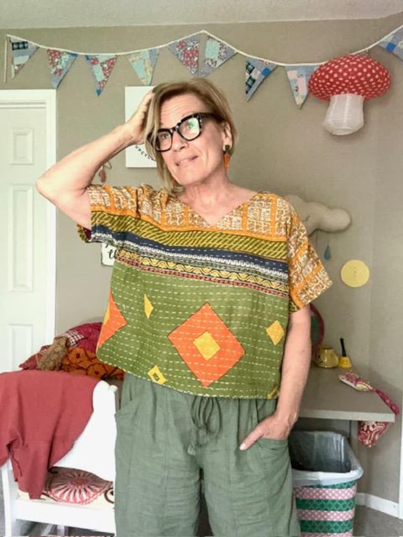 May include: A short-sleeved, boxy-fit top featuring a patchwork design with olive green, mustard yellow, and orange diamond patterns, accented by horizontal bands of dark blue and intricate white stitching throughout the fabric.