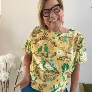 May include: A short-sleeved yellow top featuring a repeating pattern of brown line-art birdcages and baskets filled with white and blue flowers, interspersed with small green birds perched on branches and within the cages.