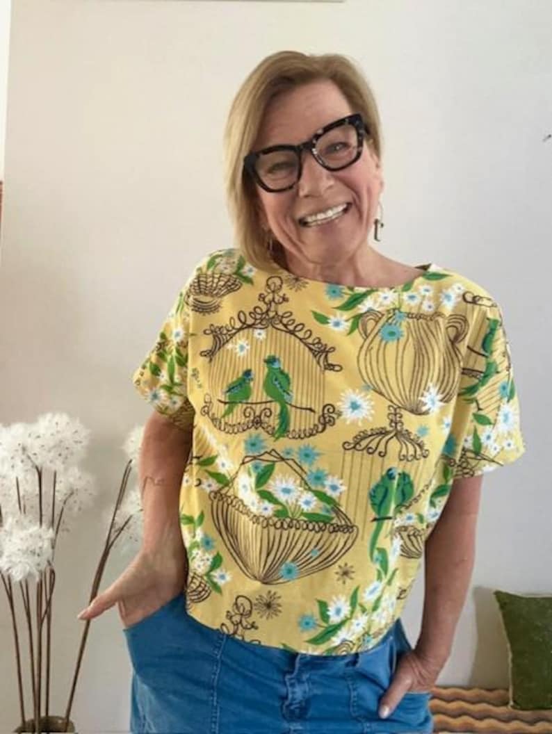 May include: A short-sleeved yellow top featuring a repeating pattern of brown line-art birdcages and baskets filled with white and blue flowers, interspersed with small green birds perched on branches and within the cages.