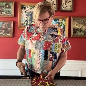May include: A short-sleeved top constructed from a patchwork of various colorful fabric scraps featuring diverse patterns, including floral, plaid, and geometric designs in shades of red, blue, yellow, and white, worn with dark-rimmed eyeglasses.