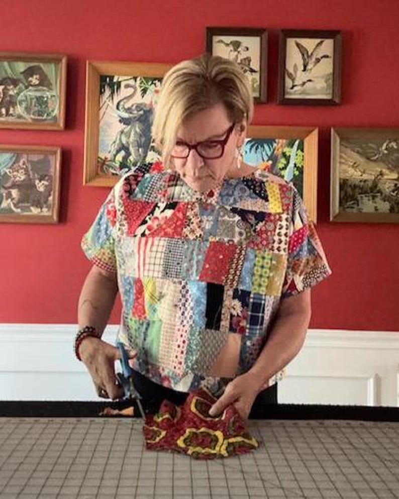 May include: A short-sleeved top constructed from a patchwork of various colorful fabric scraps featuring diverse patterns, including floral, plaid, and geometric designs in shades of red, blue, yellow, and white, worn with dark-rimmed eyeglasses.