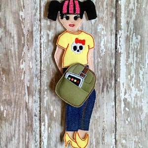 May include: A felt doll with black hair, pink and black striped bangs, a yellow shirt with a skull and bow, blue jeans, a green bag with a brown strap, and yellow shoes.