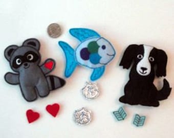 Puede incluir: Un conjunto de tres juguetes de animales de fieltro, que incluyen un mapache, un pez y un perro blanco y negro. Los juguetes están hechos a mano y tienen detalles bordados. También se incluyen corazones de fieltro, monedas y hojas.