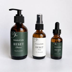 May include: Three amber glass bottles with black caps. The left bottle has a pump and a dark green label reading Botanical Belle RESET Cleanse Net 4 fl oz (118 mL). The middle bottle has a spray nozzle and white label reading Botanical Belle RESET Mist Net 2 fl oz (59 mL). The right bottle has a dropper and dark green label reading Botanical Belle RESET oil Net 1 fl oz (30 mL).