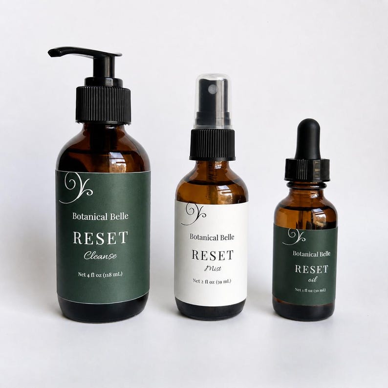 May include: Three amber glass bottles with black caps. The left bottle has a pump and a dark green label reading Botanical Belle RESET Cleanse Net 4 fl oz (118 mL). The middle bottle has a spray nozzle and white label reading Botanical Belle RESET Mist Net 2 fl oz (59 mL). The right bottle has a dropper and dark green label reading Botanical Belle RESET oil Net 1 fl oz (30 mL).