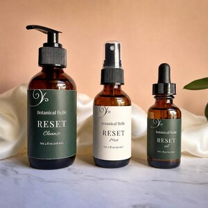 May include: Three amber glass bottles with black caps. The left bottle has a pump and a dark green label reading Botanical Belle RESET Cleanse Net 4 fl oz (118 mL). The center bottle has a spray nozzle and a white label reading Botanical Belle RESET Mist Net 2 fl oz (60 mL). The right bottle has a dropper and a dark green label reading Botanical Belle RESET oil Net 1 fl oz (30 mL).