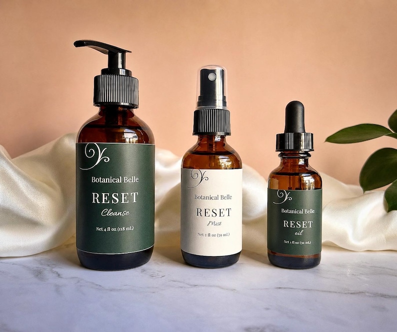 May include: Three amber glass bottles with black caps. The left bottle has a pump and a dark green label reading Botanical Belle RESET Cleanse Net 4 fl oz (118 mL). The center bottle has a spray nozzle and a white label reading Botanical Belle RESET Mist Net 2 fl oz (60 mL). The right bottle has a dropper and a dark green label reading Botanical Belle RESET oil Net 1 fl oz (30 mL).