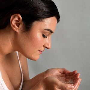 May include: A person with dark hair wearing a white tank top, cupping water in their hands to splash their face. The skin appears damp with visible water droplets, reflecting a routine of facial cleansing or refreshing with clear water.