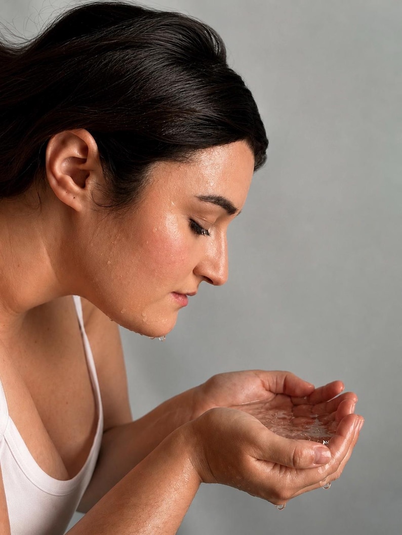 May include: A person with dark hair wearing a white tank top, cupping water in their hands to splash their face. The skin appears damp with visible water droplets, reflecting a routine of facial cleansing or refreshing with clear water.