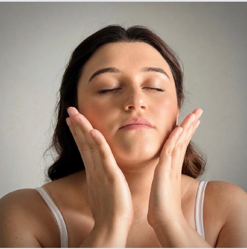 May include: A person with dark brown hair and a white tank top with thin straps, shown from the chest up. The person has their eyes closed and is gently touching both sides of their face with their hands, palms facing inward toward the cheeks.