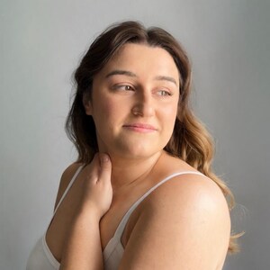 May include: A person with long, wavy brown hair and a fair complexion, wearing a simple white tank top with thin straps. The individual is looking toward the right side of the frame with a neutral expression, with one hand resting near the neck.