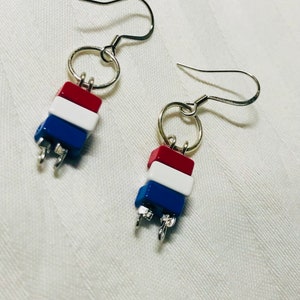 Pode incluir: Um par de brincos pendurados com contas vermelhas, brancas e azuis. Cada brinco apresenta uma pilha de três contas retangulares, conectadas por anéis e ganchos de orelha de metal prateado. Fundo branco.