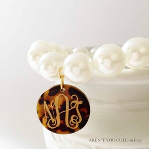 May include: A white pearl bracelet with a gold charm featuring a monogram in a tortoiseshell design.