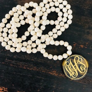 May include: A long white beaded necklace with a large, round pendant. The pendant has a tortoiseshell pattern with gold-colored initials. The beads are round and have a marbled appearance.