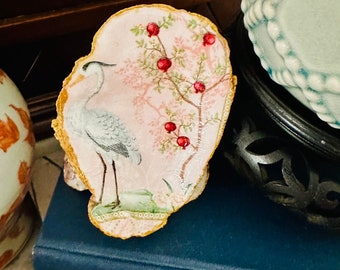 May include: A decorative oyster shell with a pink and green floral design featuring a white bird and red berries. The shell is trimmed with gold paint.