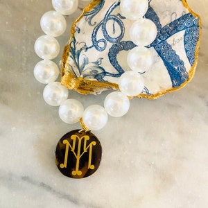 May include: A white pearl bracelet with a gold monogram charm. The charm is on a brown tortoiseshell background. The bracelet is displayed on a blue and white seashell.
