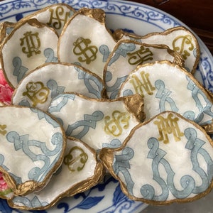 May include: A collection of white oyster shells with gold trim and blue and gold designs. Each shell has a different gold monogram.