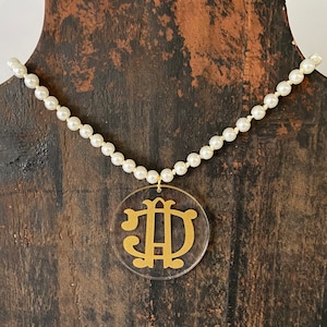 May include: A pearl necklace with a gold monogram pendant. The pendant is a circle with the letters "JD" in a script font.