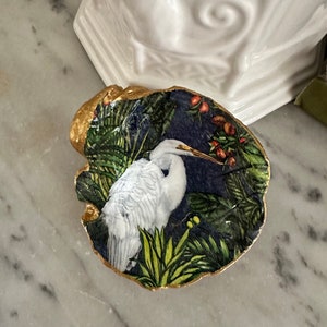 May include: A white ceramic dish with a gold rim features a painted image of a white egret standing in a lush green and blue tropical foliage.