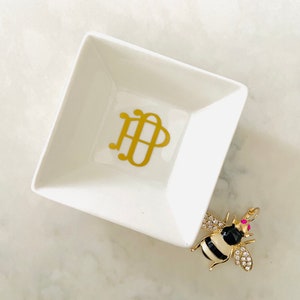 May include: White ceramic trinket dish with a gold monogram "HDP" and a gold bee charm with black and white stripes and pink accents.