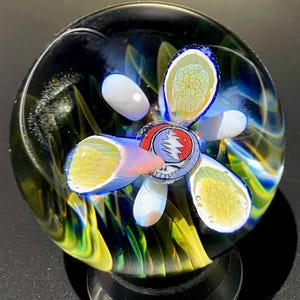 May include: A glass paperweight with a swirling blue, green, and yellow design. A white flower with a Grateful Dead logo in the center is visible inside the paperweight.