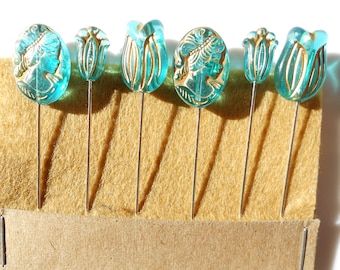 May include: Six turquoise blue glass flower head pins with gold accents. The pins are arranged in a row on a brown cardboard backing.