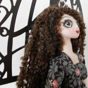 Puede incluir: Una mu&ntilde;eca con cabello casta&ntilde;o largo y rizado, rasgos faciales pintados que incluyen ojos grandes y labios rojos, vistiendo un vestido de tela oscura con un estampado floral verde y rosa. Un anillo de metal plateado est&aacute; unido a la cabeza.