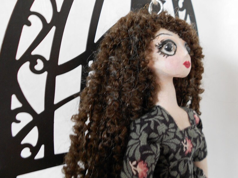 Puede incluir: Una mu&ntilde;eca con cabello casta&ntilde;o largo y rizado, rasgos faciales pintados que incluyen ojos grandes y labios rojos, vistiendo un vestido de tela oscura con un estampado floral verde y rosa. Un anillo de metal plateado est&aacute; unido a la cabeza.