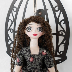 Puede incluir: Una mu&ntilde;eca con cabello largo y rizado de color marr&oacute;n, ojos grandes pintados y labios rojos, que viste un vestido gris oscuro con estampado floral. La mu&ntilde;eca est&aacute; colocada frente a un marco de metal negro decorativo en arco con una silueta de ave.