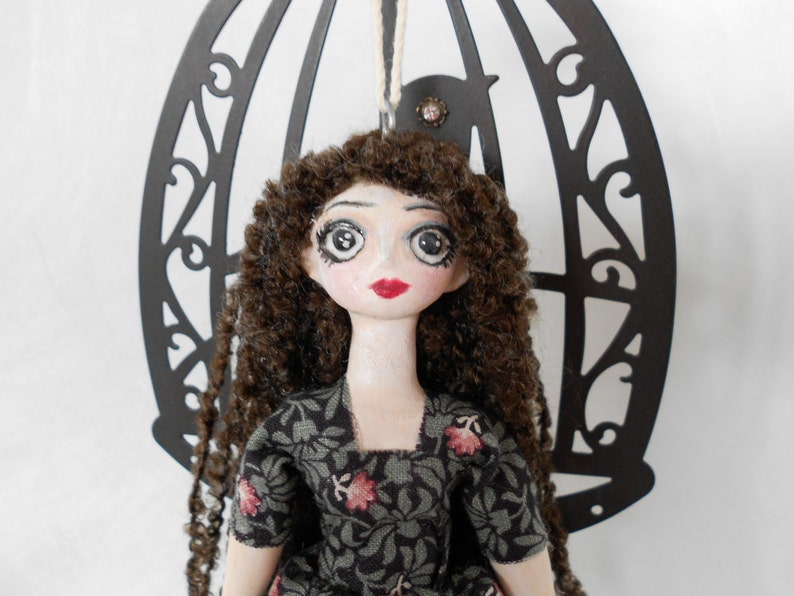 Puede incluir: Una mu&ntilde;eca con cabello largo y rizado de color marr&oacute;n, ojos grandes pintados y labios rojos, que viste un vestido gris oscuro con estampado floral. La mu&ntilde;eca est&aacute; colocada frente a un marco de metal negro decorativo en arco con una silueta de ave.
