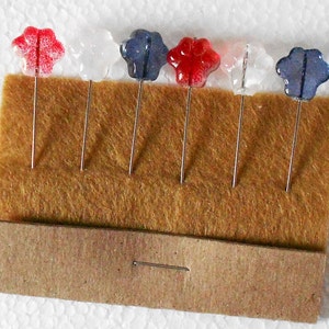 Puede incluir: Seis pequeñas alfileres de vidrio con forma de flor con detalles en rojo, blanco y azul. Los alfileres están dispuestos en una fila sobre una superficie de fieltro marrón.