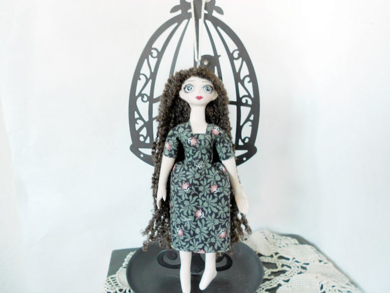 Puede incluir: Una mu&ntilde;eca de tela hecha a mano con cabello largo, rizado y casta&ntilde;o oscuro, ojos grandes azules y labios rojos. La mu&ntilde;eca viste un vestido verde oscuro con un patr&oacute;n repetitivo de hojas verdes y peque&ntilde;as flores rosas, con mangas cortas.