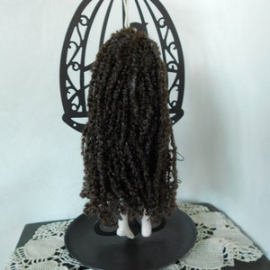Puede incluir: Una mu&ntilde;eca con cabello largo, rizado y de color marr&oacute;n oscuro que cae por la espalda, colocada frente a un soporte de metal negro con forma de jaula. La mu&ntilde;eca tiene pies blancos visibles y cuelga de la parte superior de la estructura.
