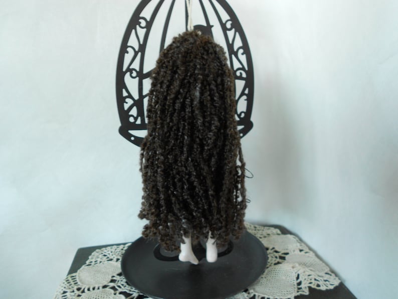 Puede incluir: Una mu&ntilde;eca con cabello largo, rizado y de color marr&oacute;n oscuro que cae por la espalda, colocada frente a un soporte de metal negro con forma de jaula. La mu&ntilde;eca tiene pies blancos visibles y cuelga de la parte superior de la estructura.