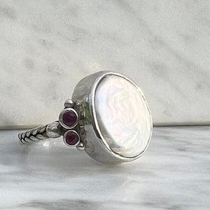May include: A silver ring with a large white pearl cabochon and three small red gemstones.