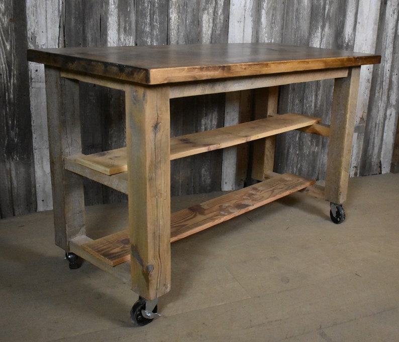 May include: Rustic wooden kitchen island with a distressed brown finish. The island has two shelves and is on wheels.