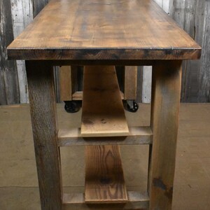 May include: A rustic wooden table with a brown stained top and three shelves. The table has four black wheels and is made of reclaimed wood.