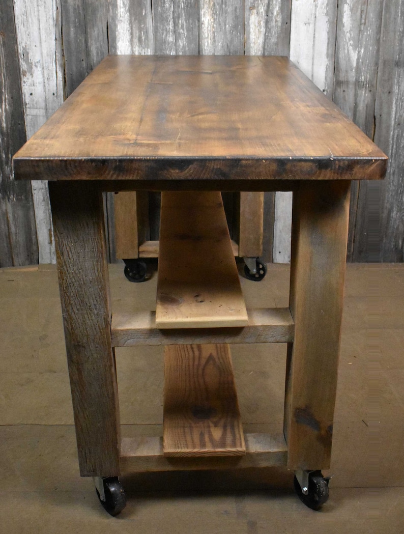 May include: A rustic wooden table with a brown stained top and three shelves. The table has four black wheels and is made of reclaimed wood.