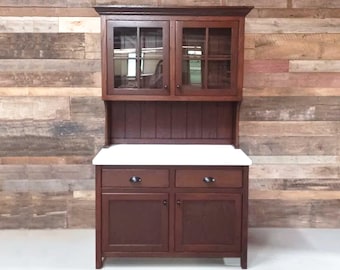 May include: A dark brown wooden hutch with glass doors and a white countertop. The hutch has two drawers and two doors with a paneled design. The hutch is against a rustic wood wall.
