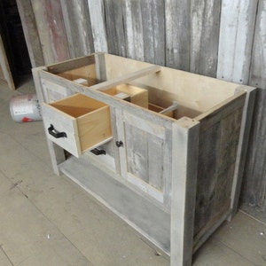 May include: A rustic wooden bathroom vanity with a single drawer and a cabinet door. The vanity is made of reclaimed wood and has a grey finish. The drawer and cabinet door have black metal handles.