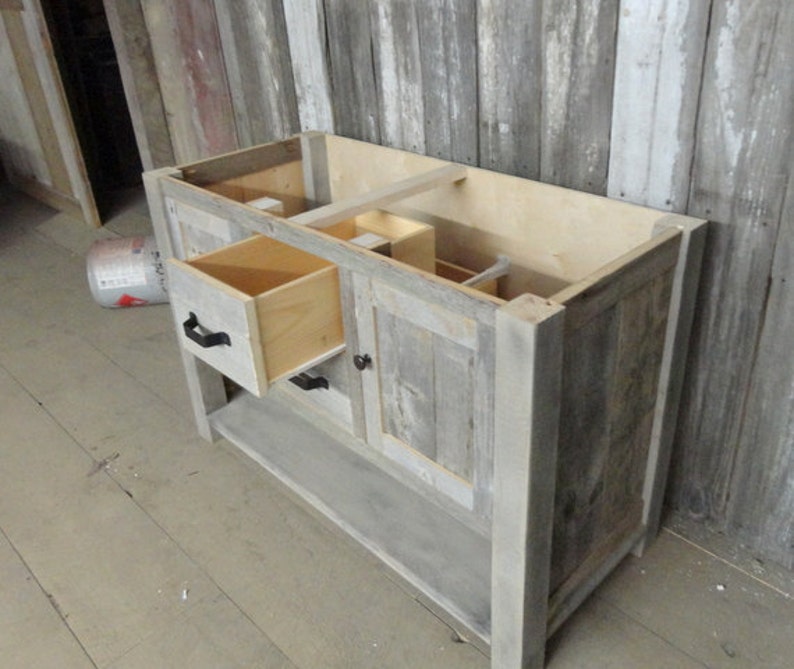 May include: A rustic wooden bathroom vanity with a single drawer and a cabinet door. The vanity is made of reclaimed wood and has a grey finish. The drawer and cabinet door have black metal handles.