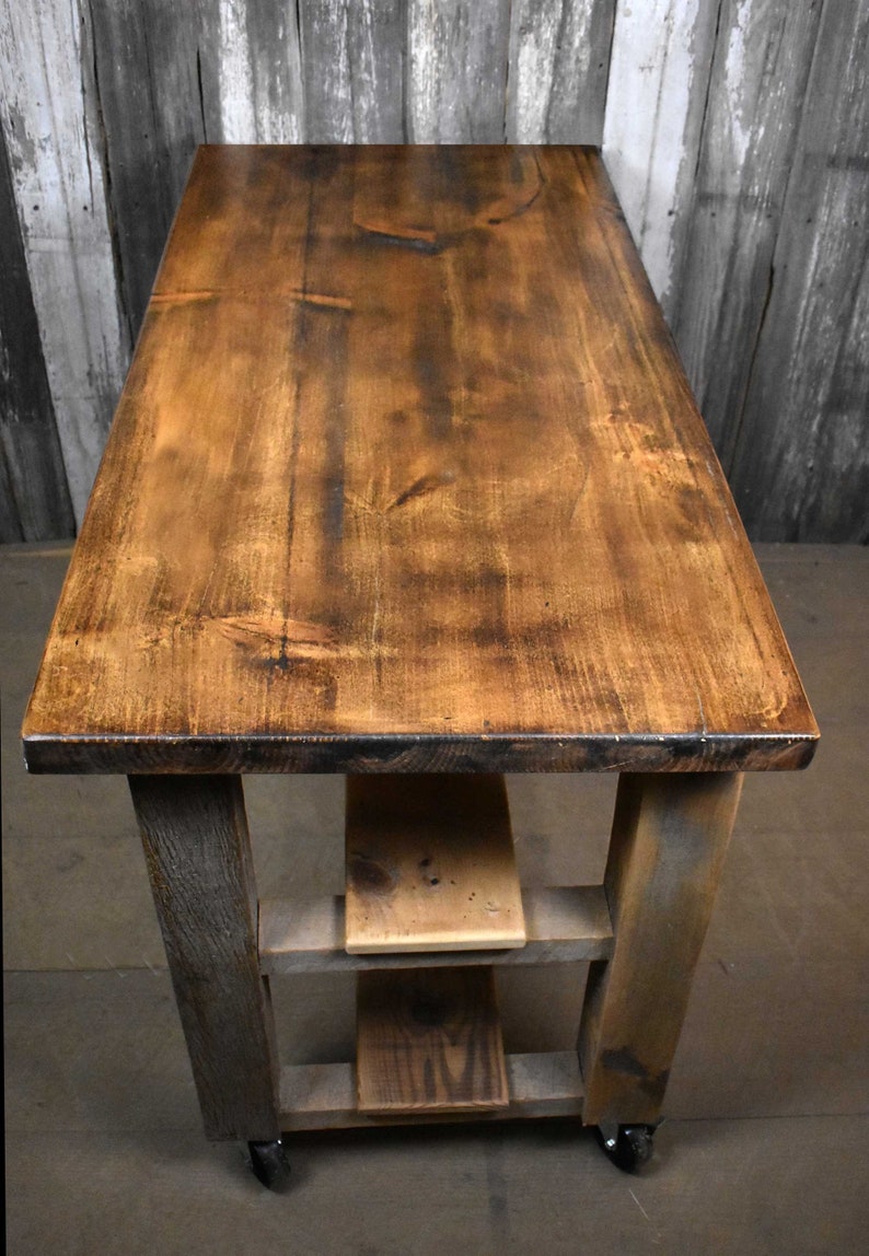 May include: A rustic wooden kitchen island with a distressed brown top and two shelves. The island is on wheels and has a gray-stained base.